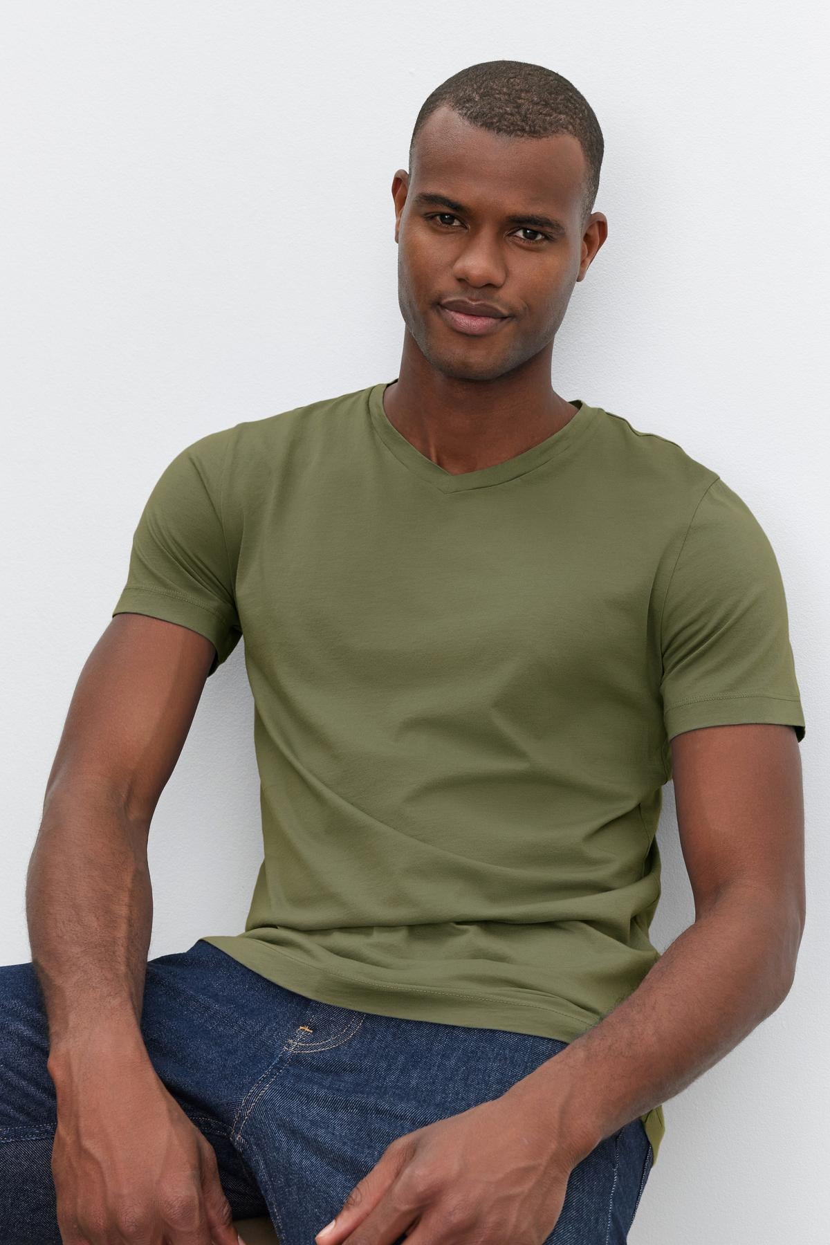   A person is wearing a green SAMSEN TEE by Velvet by Graham & Spencer and blue jeans, sitting against a light gray background. 