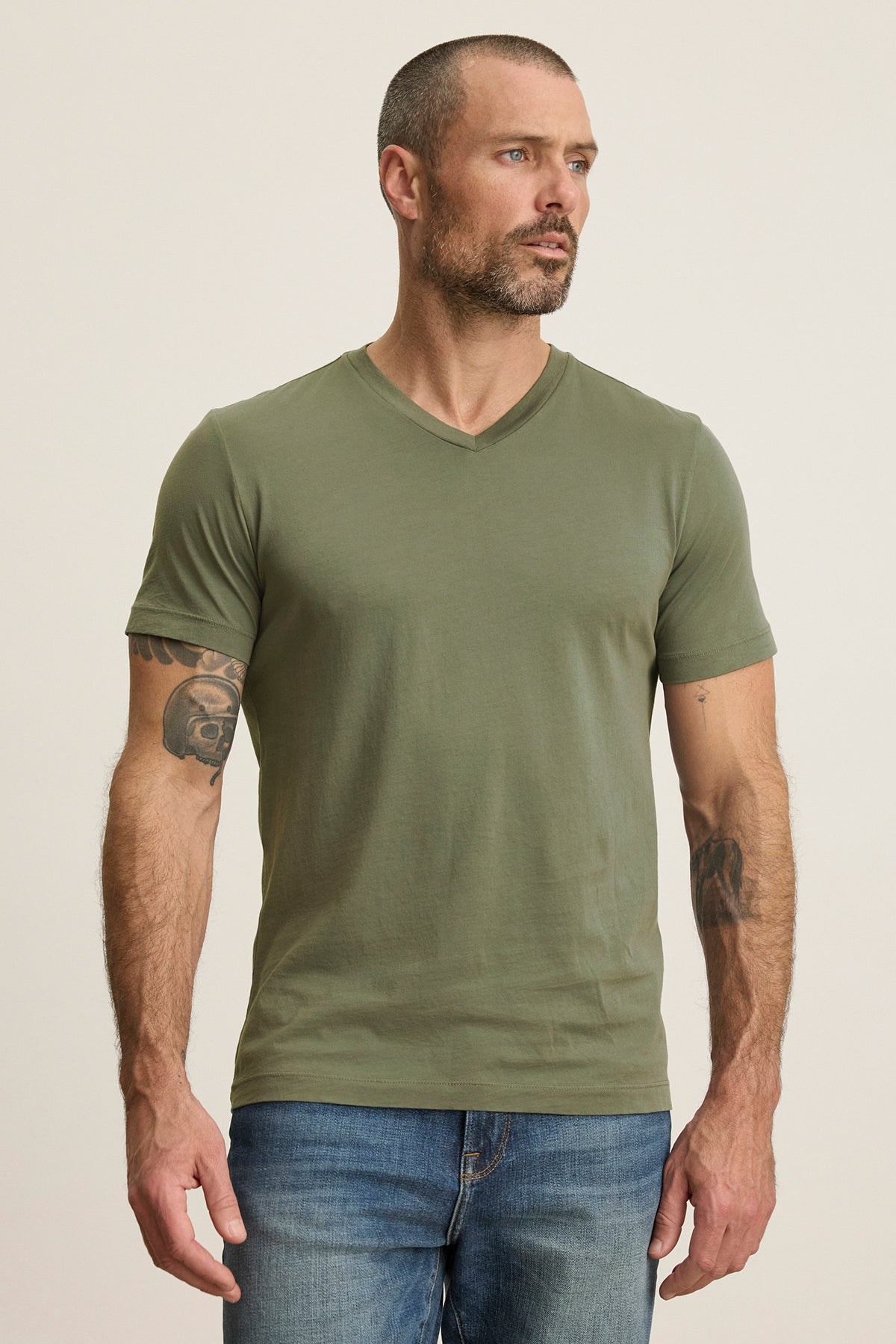   A man with a beard stands sideways, revealing tattoos on his left arm, wearing the green SAMSEN TEE by Velvet by Graham & Spencer paired with jeans. The v-neckline adds style. 