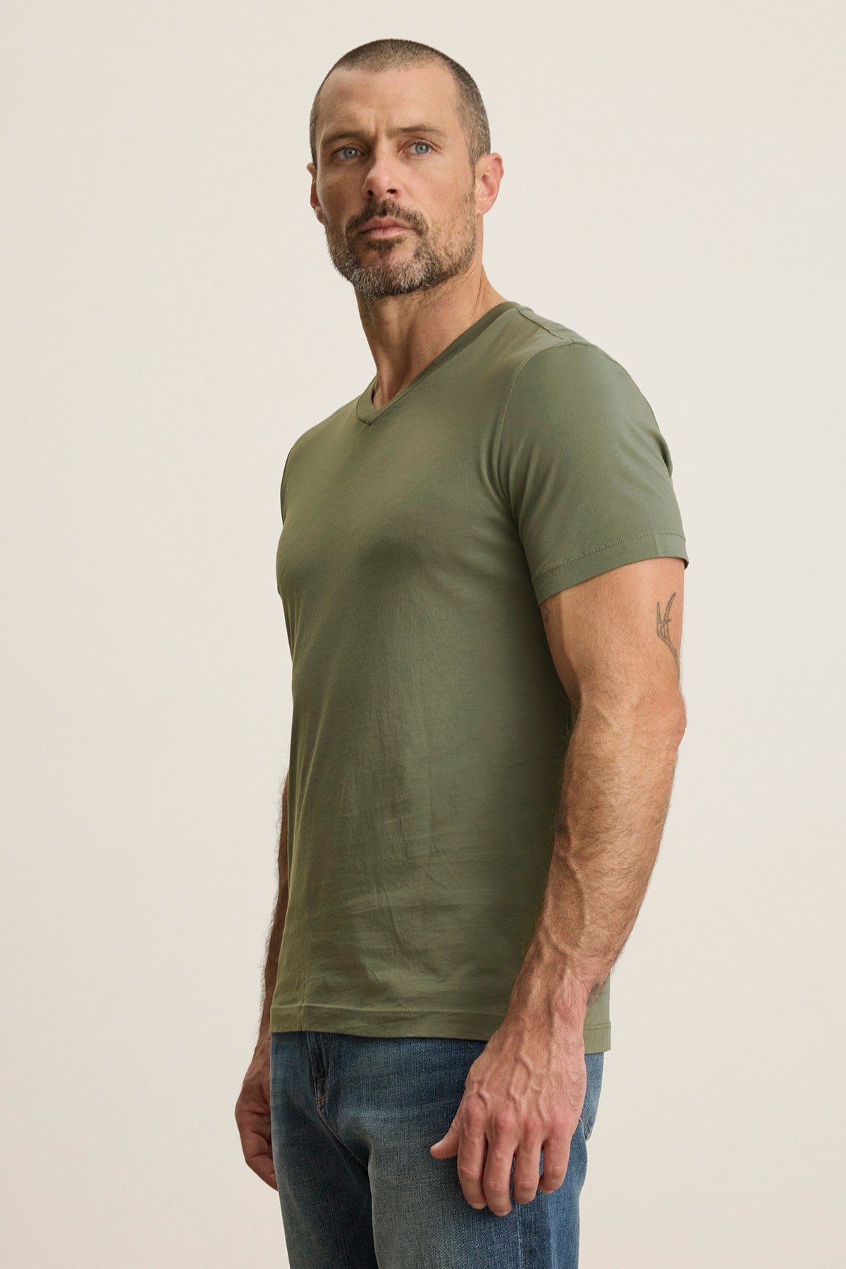 A man in a SAMSEN TEE by Velvet by Graham & Spencer, featuring a soft green Pima jersey with a v-neckline, stands against a neutral background wearing jeans and gazes slightly to the left.-38866102288577