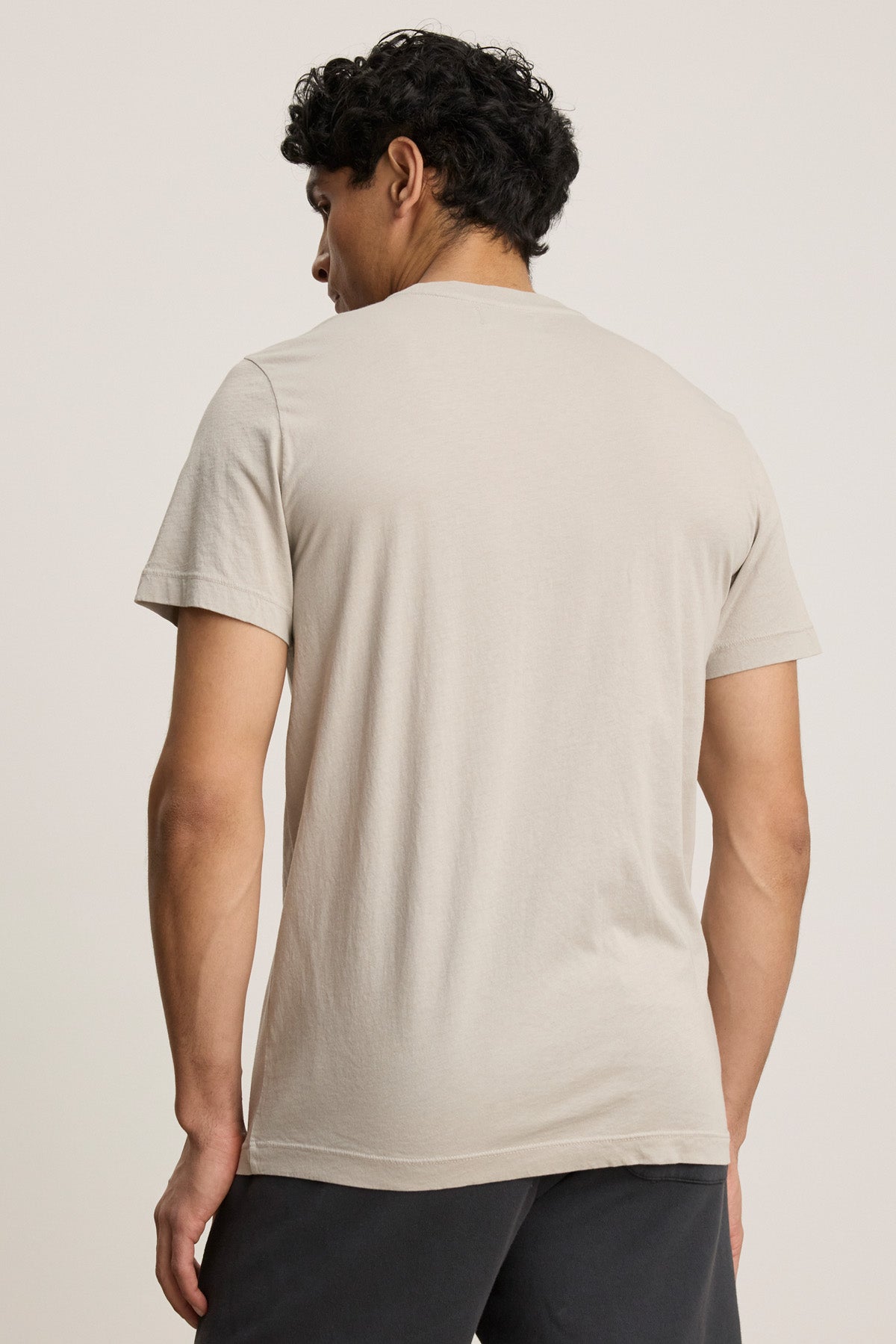 A person faces away from the camera, wearing the SAMSEN TEE by Velvet by Graham & Spencer—a light beige Pima jersey short-sleeve t-shirt—paired with dark pants against a plain background, perfect for everyday style.-40606051139777