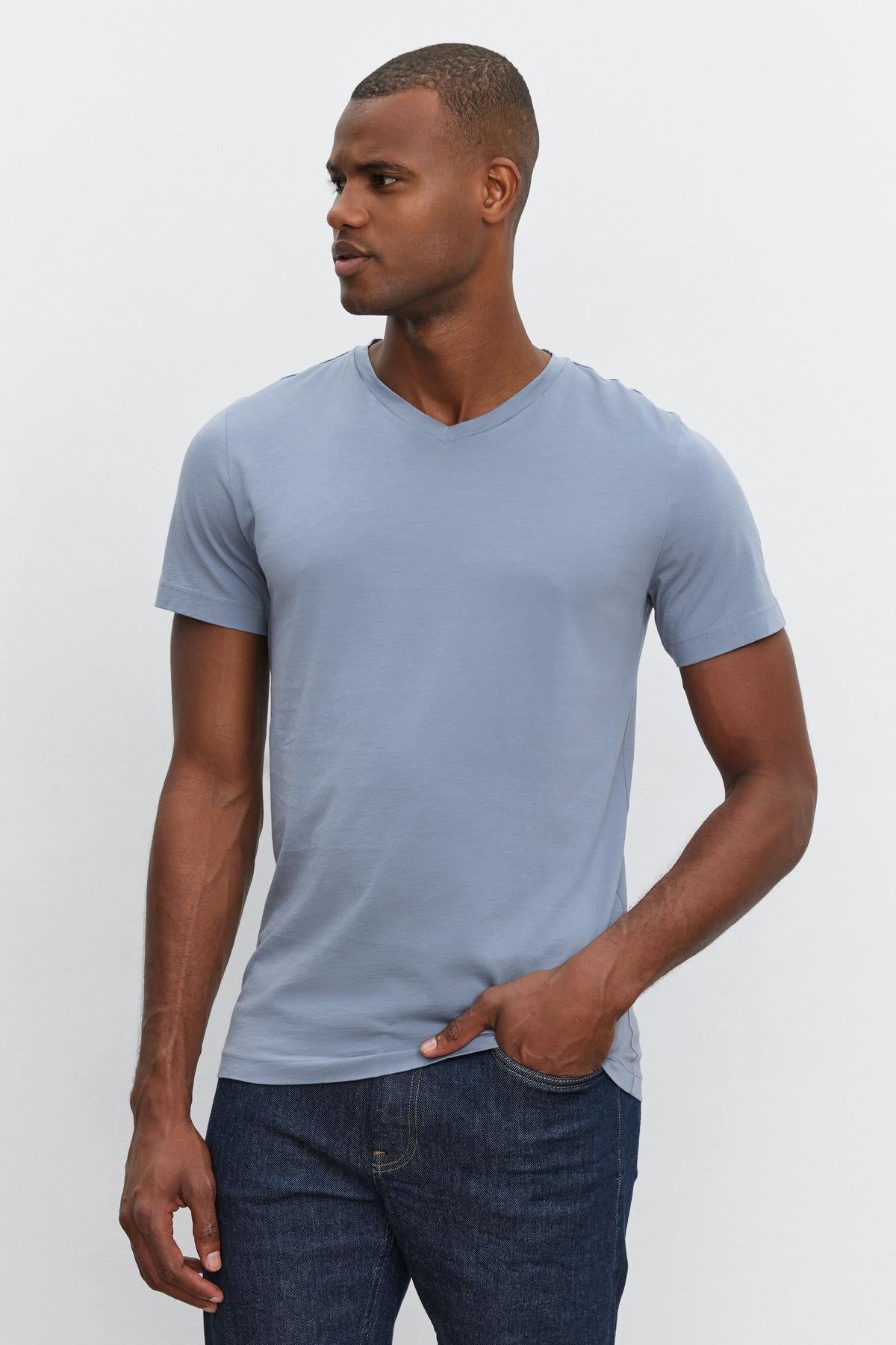 A man wearing a light blue SAMSEN TEE by Velvet by Graham & Spencer made from whisper cotton knit and dark blue jeans stands against a plain white background, looking to his left.-37386148774081