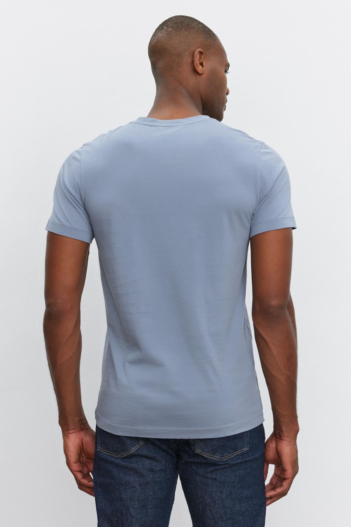 A man, clad in everyday wear— a light blue short-sleeve SAMSEN TEE made of whisper cotton knit by Velvet by Graham & Spencer and dark jeans, faces away from the camera.-37386148806849