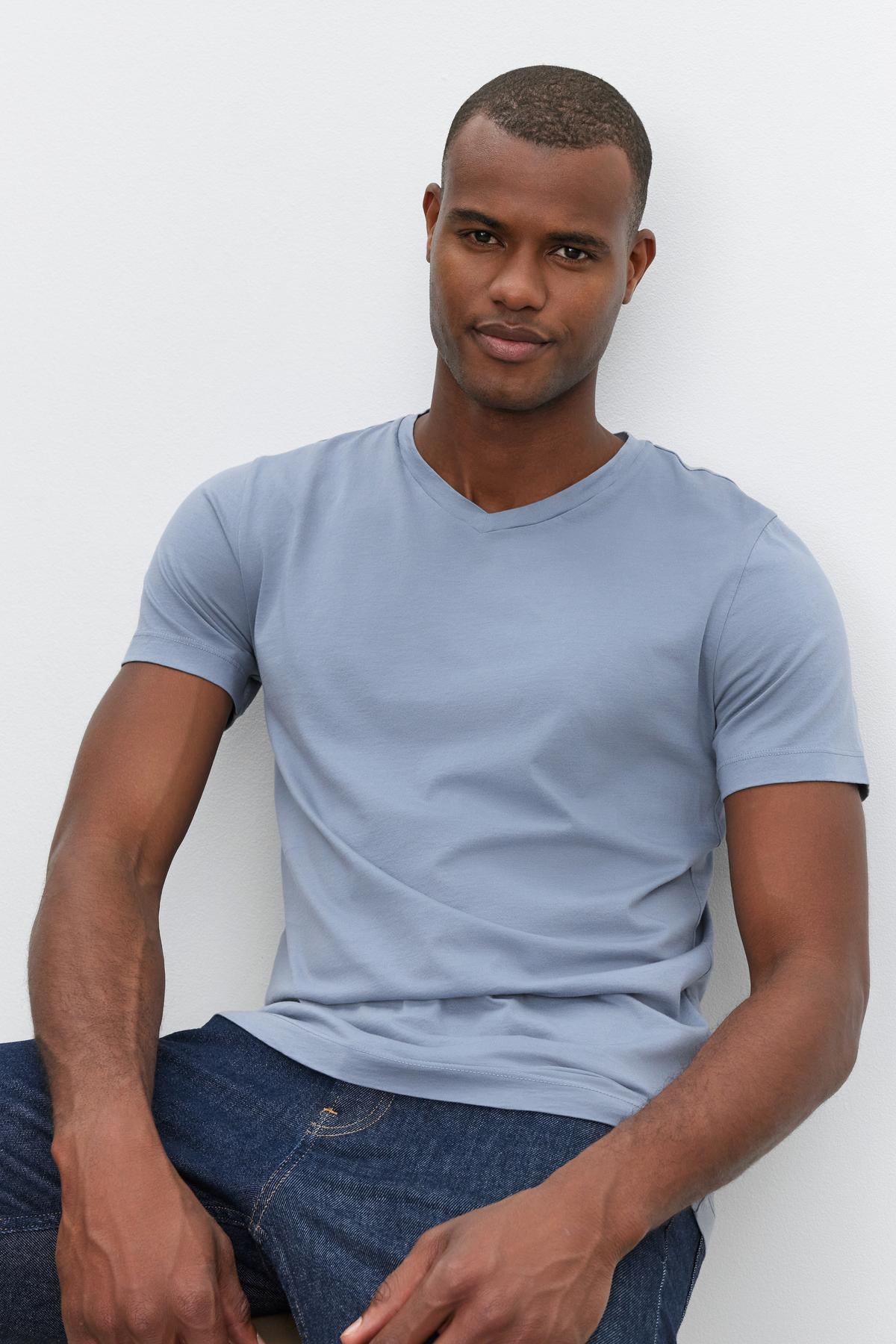A man in a light blue SAMSEN TEE by Velvet by Graham & Spencer with a whisper cotton knit and dark jeans is seated against a plain white background.-37386148708545