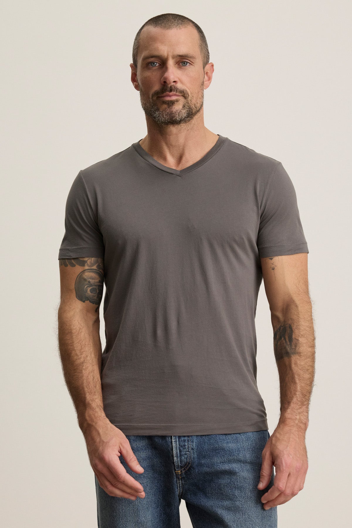 A man with short hair and a beard stands facing forward, wearing the gray SAMSEN TEE by Velvet by Graham & Spencer with blue jeans against a plain background—a versatile V-neck ideal for layering or wearing solo.-40452491608257