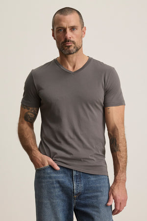 A man with short hair and a beard wears the SAMSEN TEE by Velvet by Graham & Spencer in grey, paired with blue jeans. He stands against a plain light background, hand in pocket—showcasing a relaxed yet refined style perfect for effortless layering.