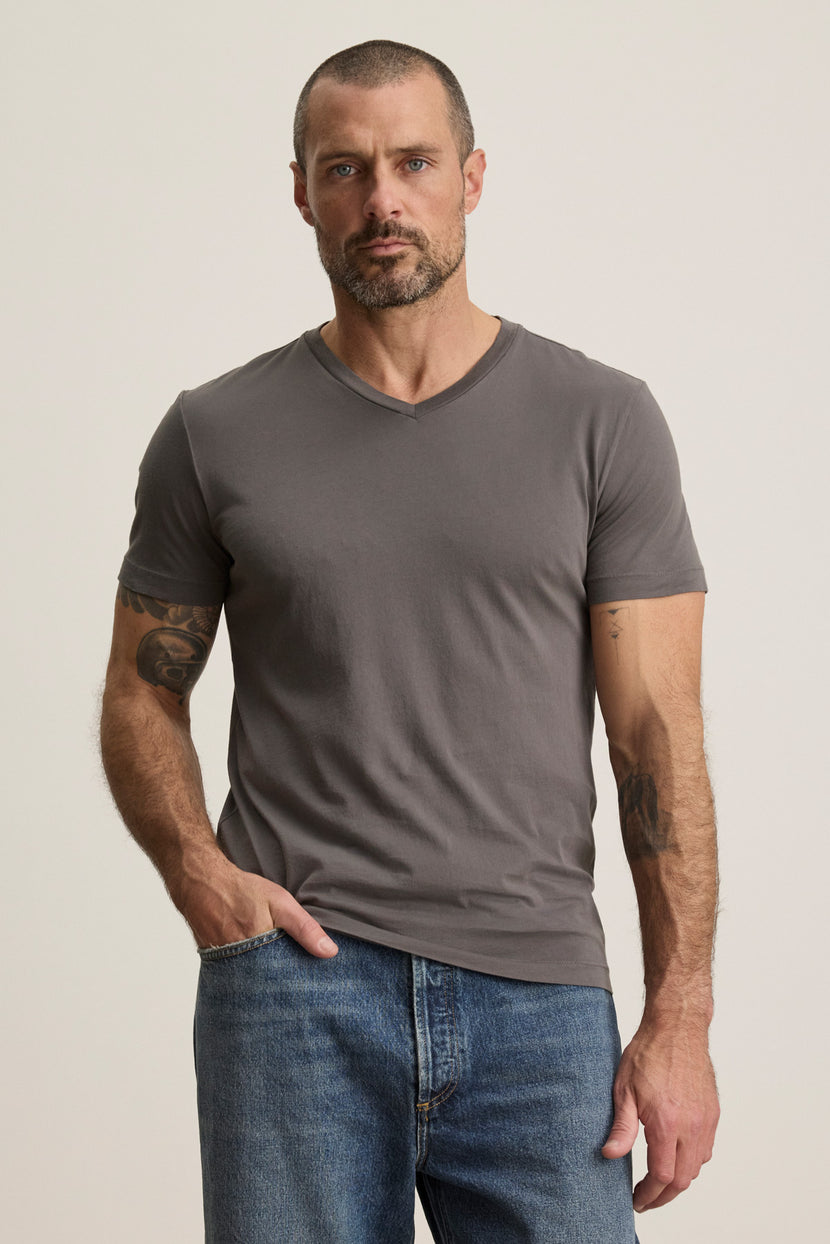 A man with short hair and a beard wears the SAMSEN TEE by Velvet by Graham & Spencer in grey, paired with blue jeans. He stands against a plain light background, hand in pocket—showcasing a relaxed yet refined style perfect for effortless layering.