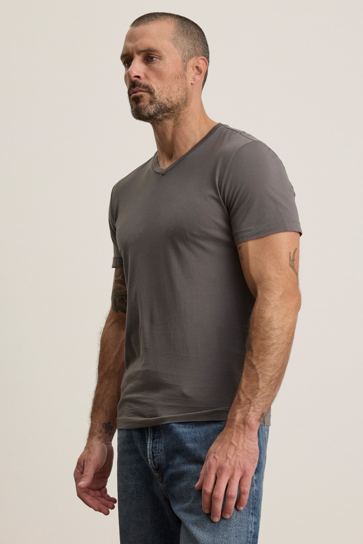Man with short hair and beard wears the SAMSEN TEE by Velvet by Graham & Spencer—a grey Pima jersey v-neck tee—paired with blue jeans against a neutral background, ideal for layering any season.-40452491542721