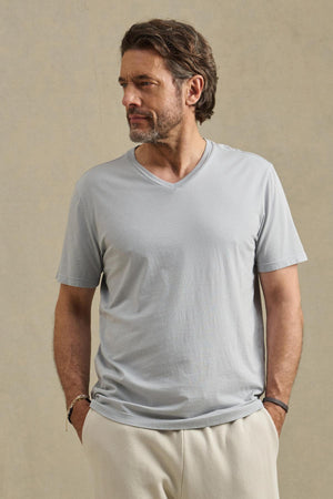 A man with short brown hair and a beard wears the SAMSEN TEE by Velvet by Graham & Spencer, a light gray Pima jersey V-neck slim-fit shirt, paired with light pants. He stands hands in pockets, looking to the side against a neutral background.