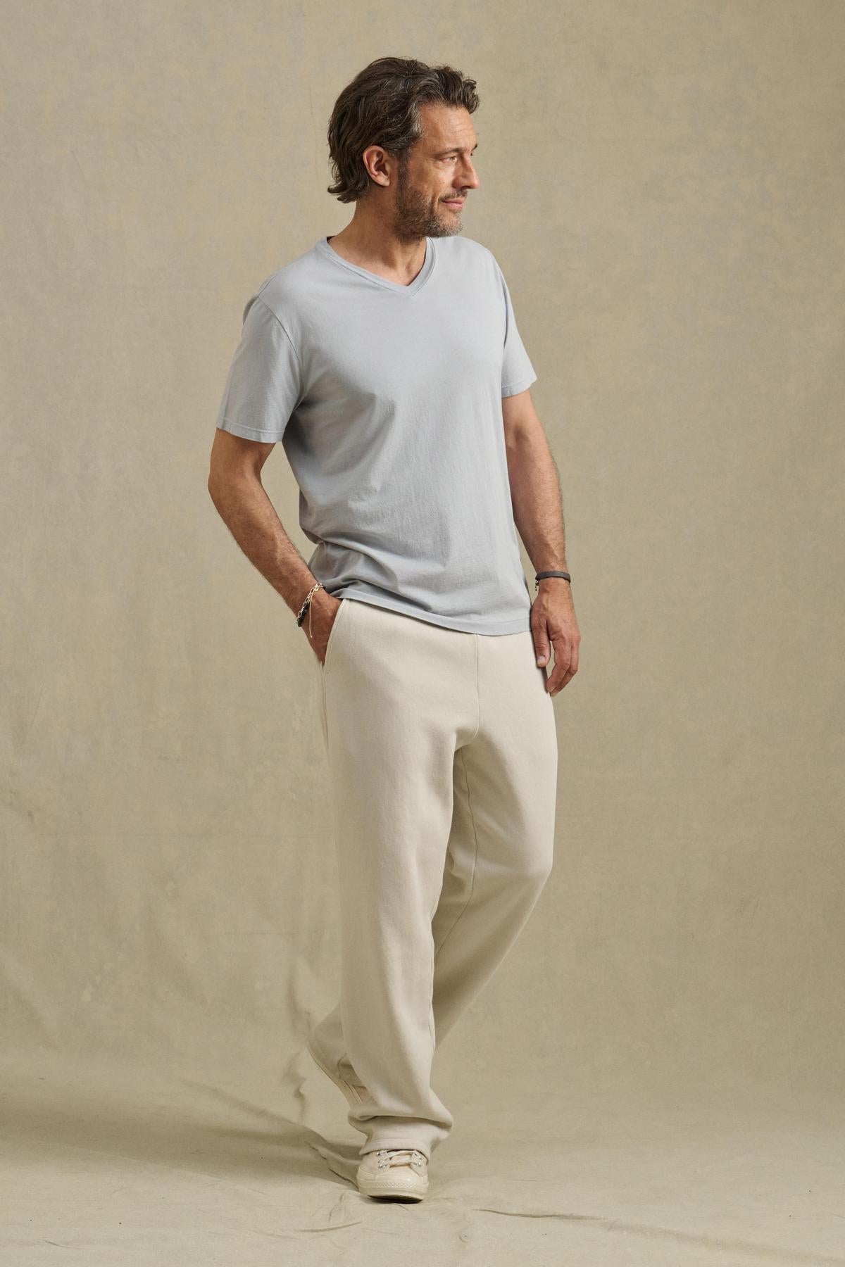 A man stands sideways against a beige background, wearing the Velvet by Graham & Spencer SAMSEN TEE in light gray, cream pants, and white sneakers.