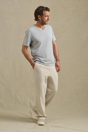 A man stands sideways against a beige background, wearing the Velvet by Graham & Spencer SAMSEN TEE in light gray, cream pants, and white sneakers.