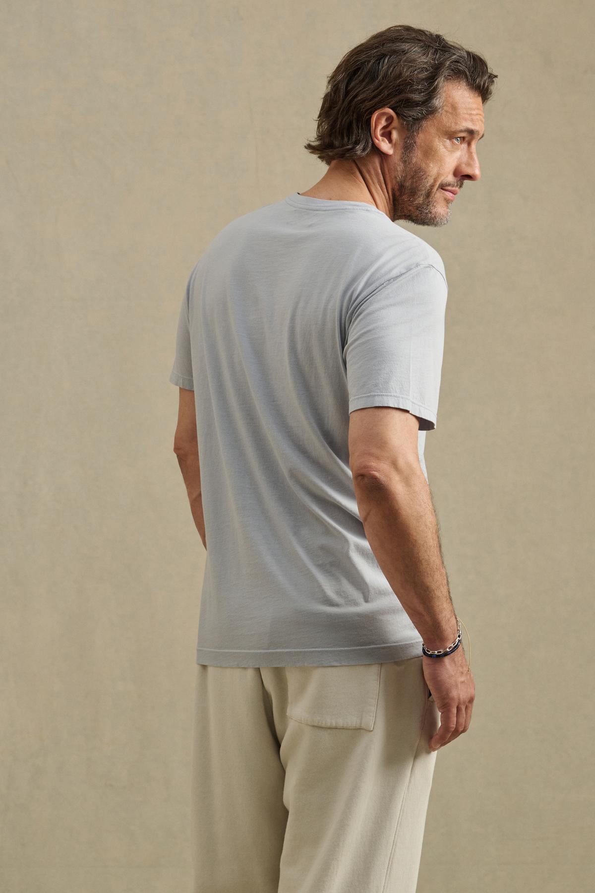 A man with brown hair and a short beard faces away, wearing the slim fit SAMSEN TEE by Velvet by Graham & Spencer in light gray with beige pants, standing against a plain beige background.-41453747863745