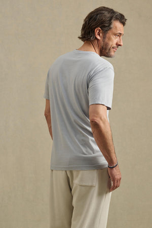 A man with brown hair and a short beard faces away, wearing the slim fit SAMSEN TEE by Velvet by Graham & Spencer in light gray with beige pants, standing against a plain beige background.