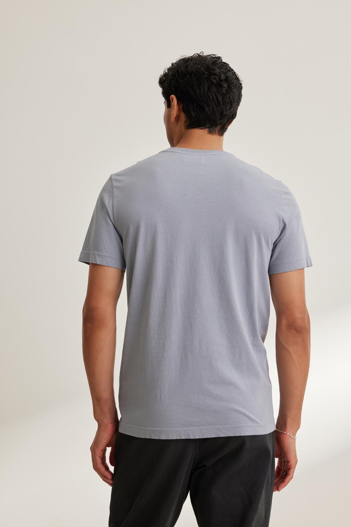 A person, with their back to the camera, wears a Velvet by Graham & Spencer SAMSEN TEE in light gray Pima jersey featuring a v-neckline, paired with dark pants against a plain background.-38643347194049