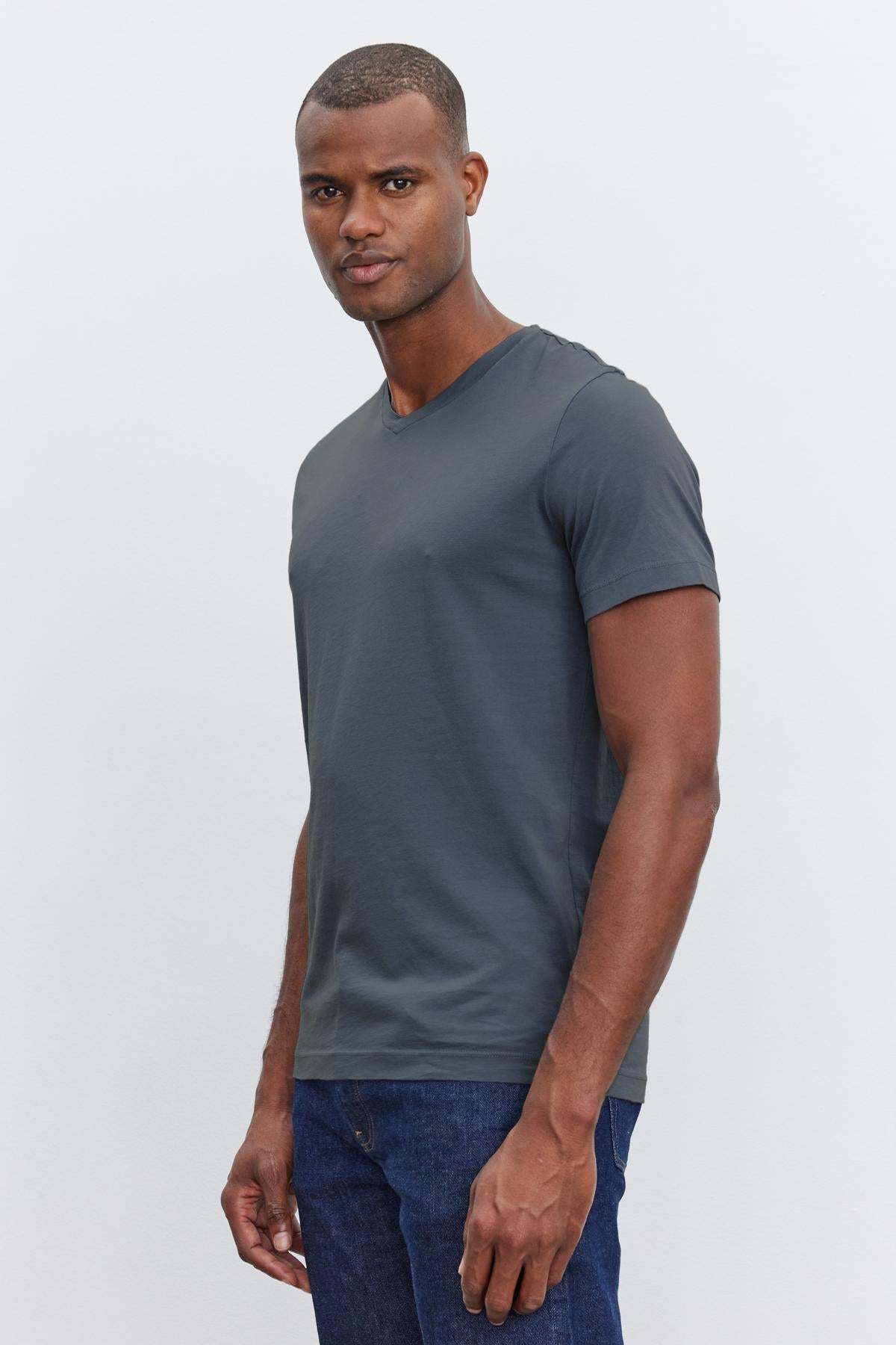 A man wearing a dark gray short-sleeve, v-neckline SAMSEN TEE by Velvet by Graham & Spencer and blue jeans stands against a plain light background. He is looking directly at the camera, embodying effortless everyday wear.-37386135437505
