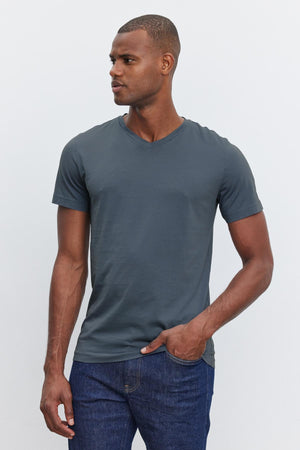 A man, showcasing everyday wear, is dressed in a fitted dark gray V-neck SAMSEN TEE by Velvet by Graham & Spencer and blue jeans. He stands against a plain light background, looking to the side with one hand in his pocket.