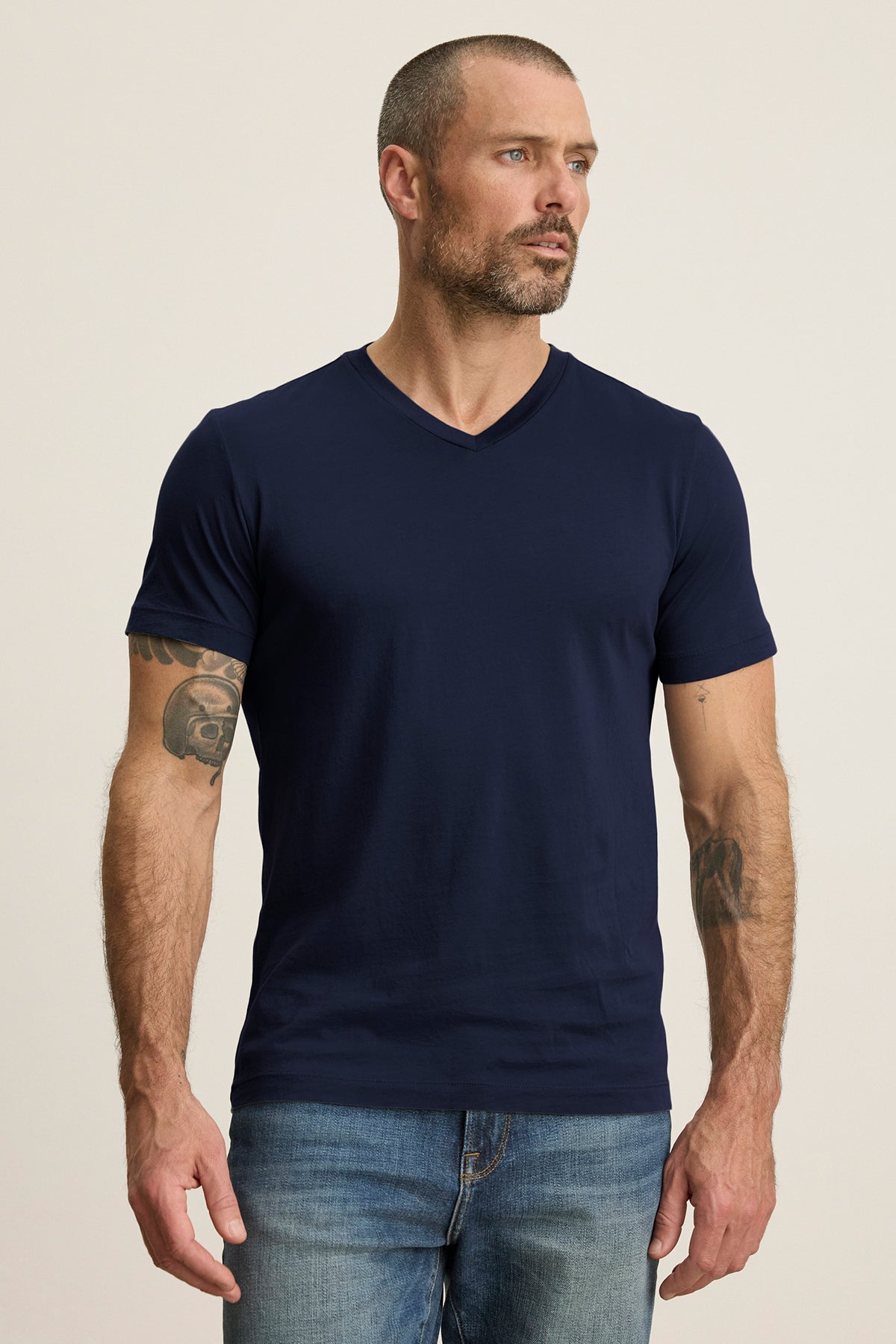 A bearded man in a Velvet by Graham & Spencer SAMSEN TEE, featuring a navy blue V-neckline Pima jersey, stands with hands relaxed at his sides while wearing jeans.-38866076958913