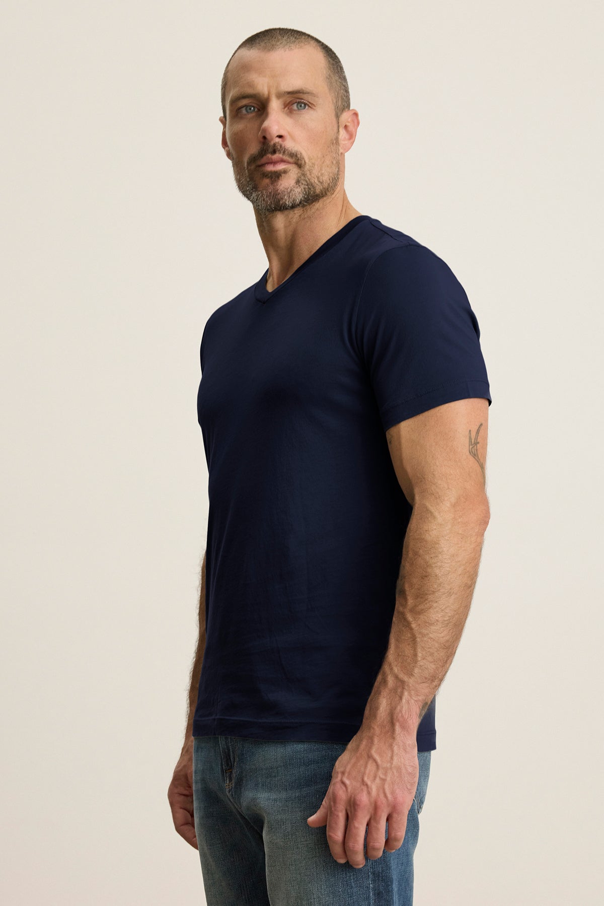   A man with a beard stands against a plain background, wearing a SAMSEN TEE from Velvet by Graham & Spencer, a navy v-neck crafted from soft Pima jersey, paired with jeans. 