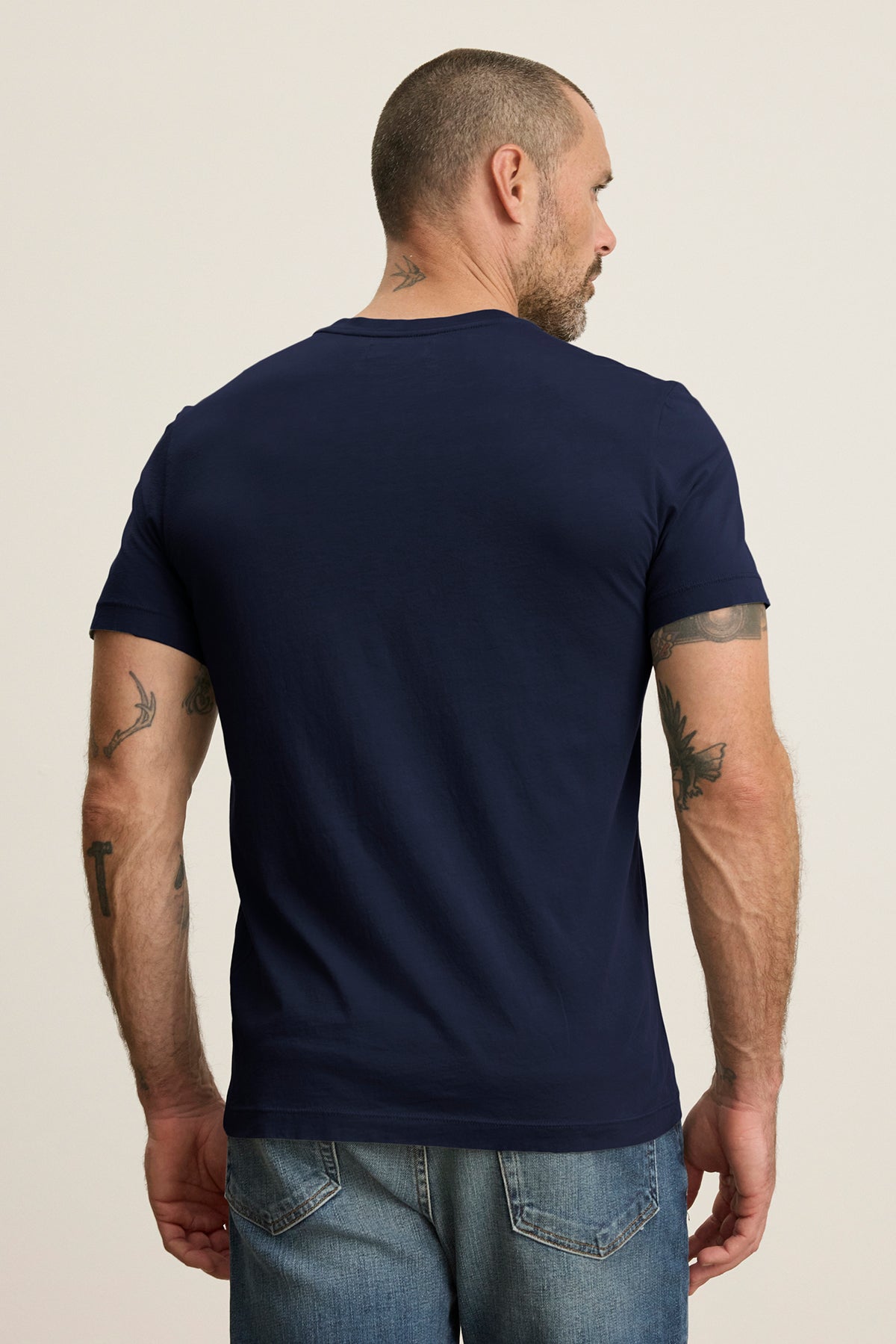 A man with short hair and tattoos on his arms and neck wears the SAMSEN TEE by Velvet by Graham & Spencer, a navy blue Pima jersey with a v-neckline, paired with jeans as he looks away from the camera.-38866076991681