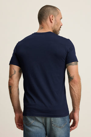 A man with short hair and tattoos on his arms and neck wears the SAMSEN TEE by Velvet by Graham & Spencer, a navy blue Pima jersey with a v-neckline, paired with jeans as he looks away from the camera.