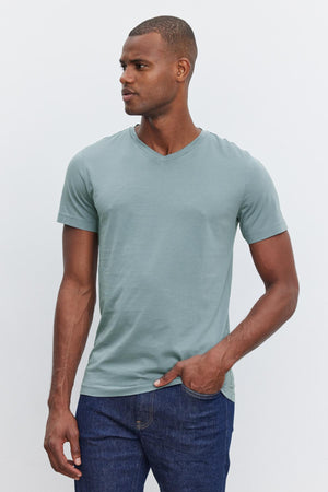 A man wearing a light green SAMSEN TEE made of whisper cotton knit by Velvet by Graham & Spencer and dark blue jeans stands against a plain light-colored background, looking to the side. This ensemble showcases stylish everyday wear with its comfortable fit and modern v-neckline.