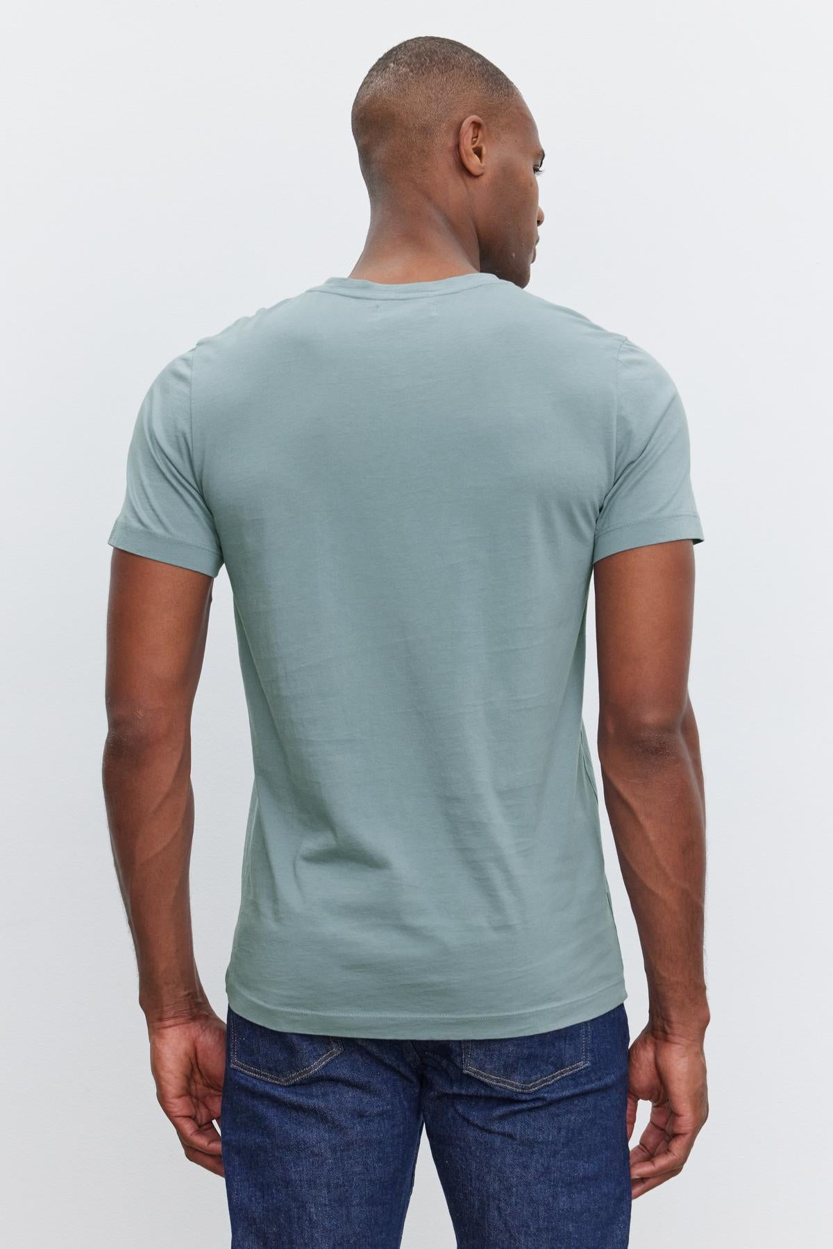   A person is standing with their back to the camera, wearing a light blue SAMSEN TEE by Velvet by Graham & Spencer and dark blue jeans against a plain white background. 