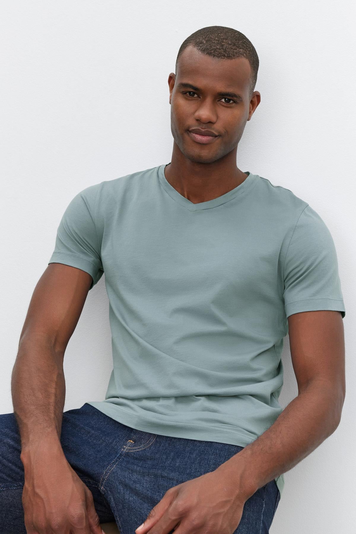   A man wearing a light green SAMSEN TEE by Velvet by Graham & Spencer with a v-neckline and dark blue jeans poses sitting against a plain white background, perfect for everyday wear. 