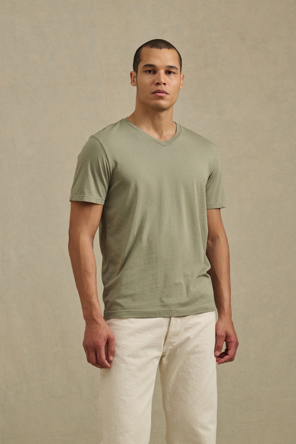 A man stands before a beige backdrop in the Velvet by Graham & Spencer SAMSEN TEE, a light olive green Pima jersey with a V-neckline, paired with off-white pants—an ideal everyday look.-41297105092801