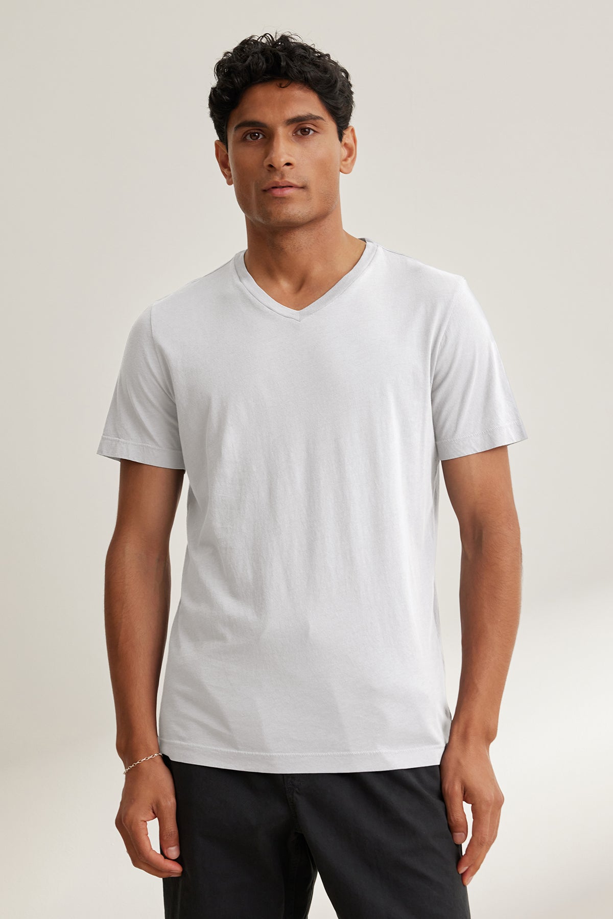   A person models the SAMSEN TEE by Velvet by Graham & Spencer, a plain white V-neck crafted from soft Pima jersey, paired with black pants, perfect for everyday wear against a neutral backdrop. 