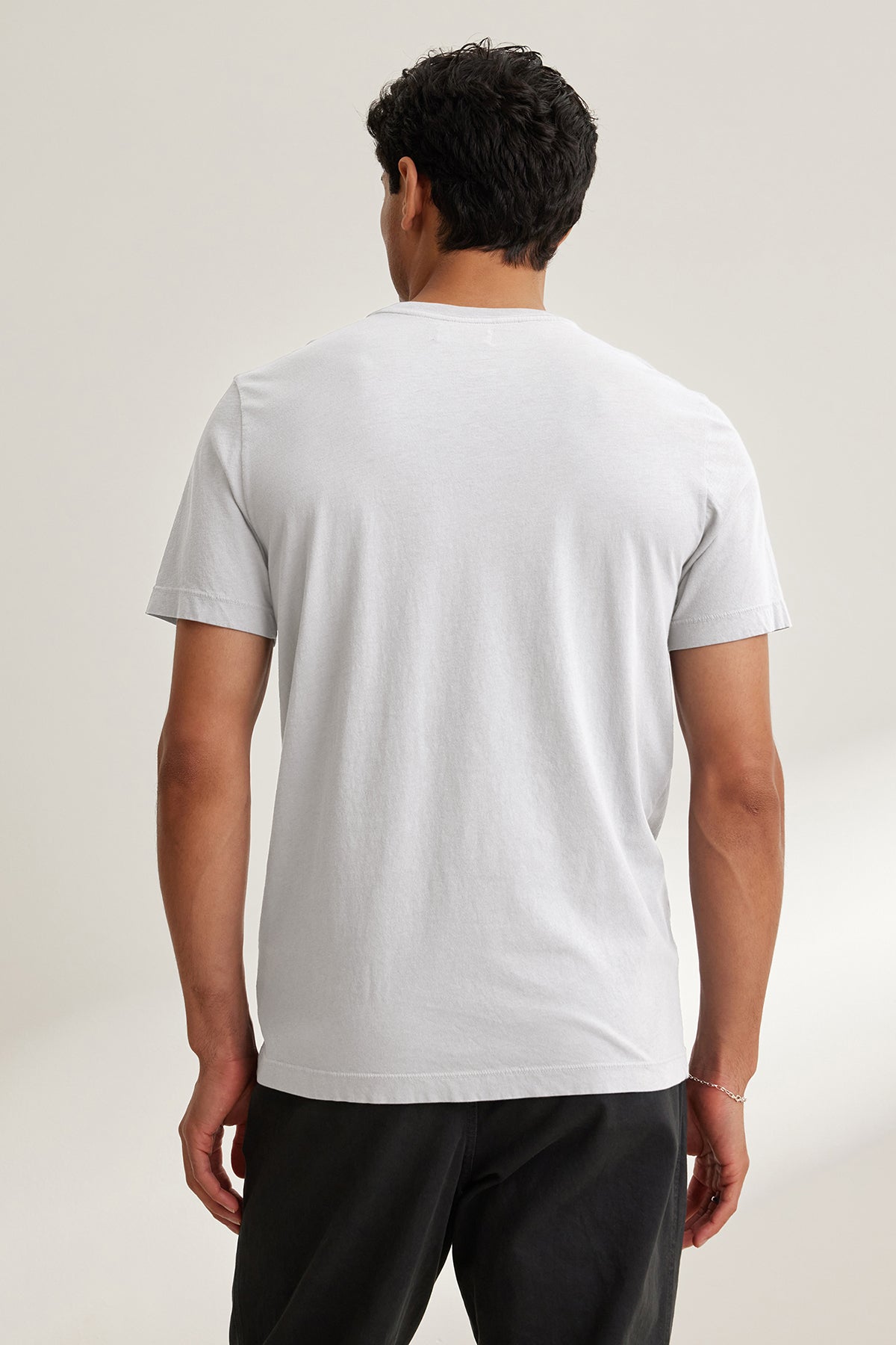   A person stands with their back to the camera, wearing a SAMSEN TEE by Velvet by Graham & Spencer—a plain white T-shirt with a v-neckline—paired with dark pants for an everyday look. 