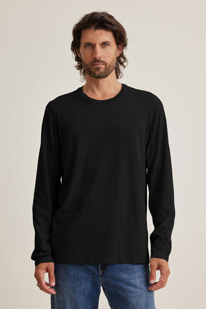 A man with wavy brown hair wears a plain black SKEETER TEE by Velvet by Graham & Spencer, crafted from soft Pima jersey, paired with blue jeans against a neutral background—perfect for any versatile wardrobe.