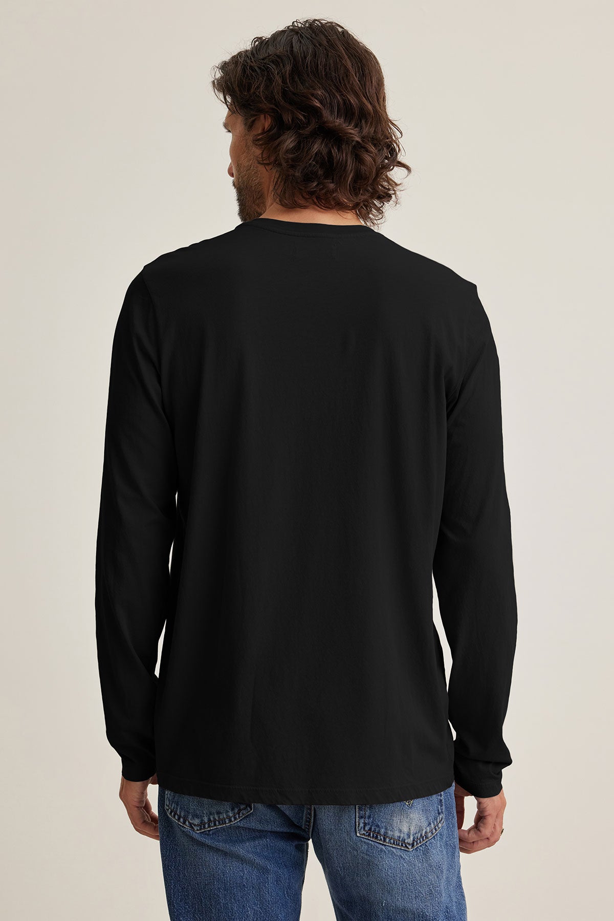 A person with wavy brown hair is seen from the back in a long-sleeve black SKEETER TEE by Velvet by Graham & Spencer and blue jeans, standing against a plain light background—a stylish pick for any wardrobe.-39144962982081