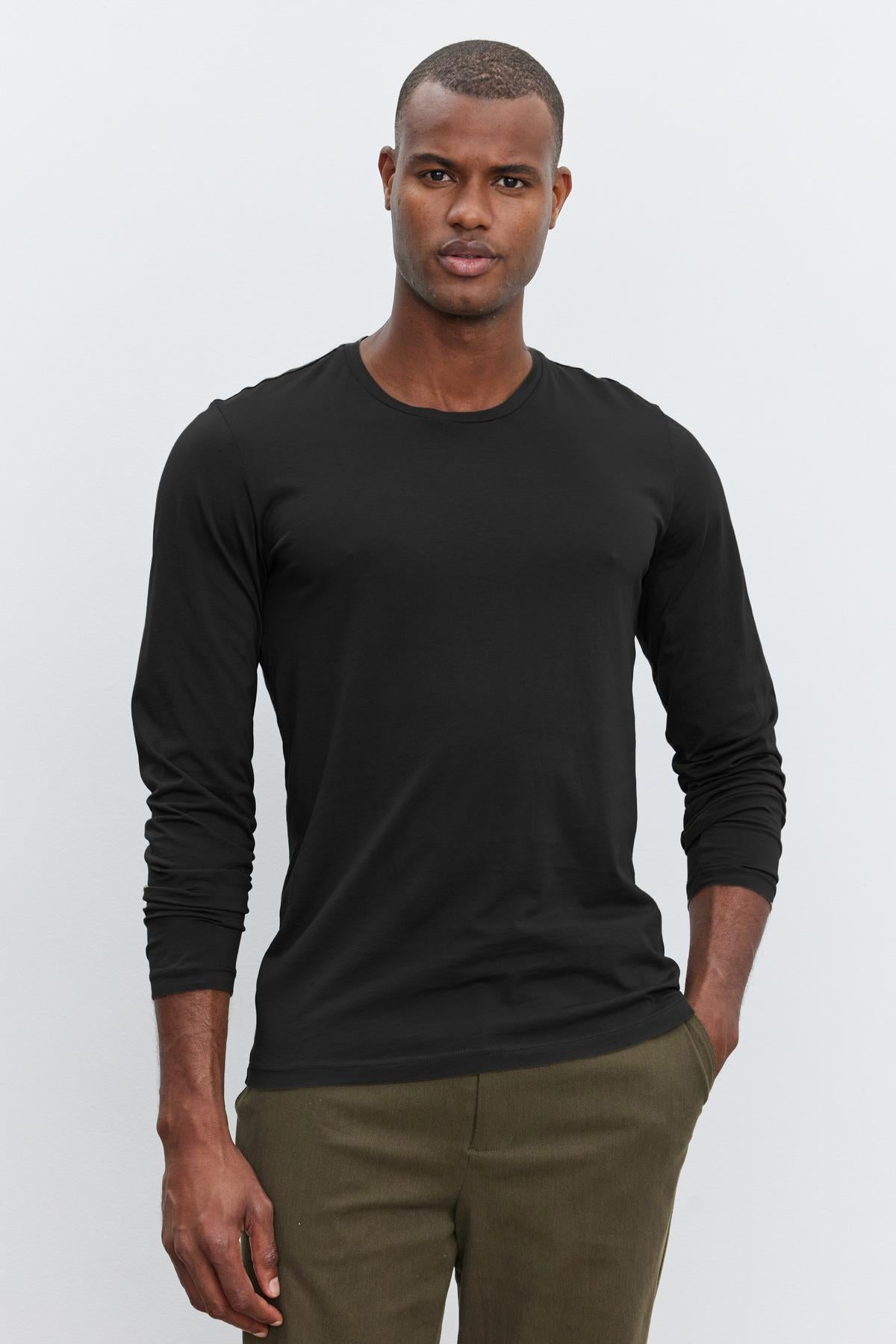   A young man in a black Velvet by Graham & Spencer SKEETER WHISPER CLASSIC CREW NECK TEE and olive green pants, standing against a white background. 