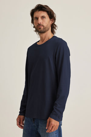 A man with medium-length brown hair and a beard wears the Velvet by Graham & Spencer SKEETER TEE in navy with blue jeans, standing against a plain light background.