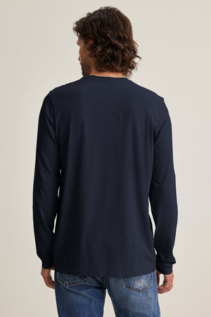 A person with medium-length brown hair stands with their back to the camera, wearing the Velvet by Graham & Spencer SKEETER TEE in soft Pima jersey and blue jeans against a plain off-white background.