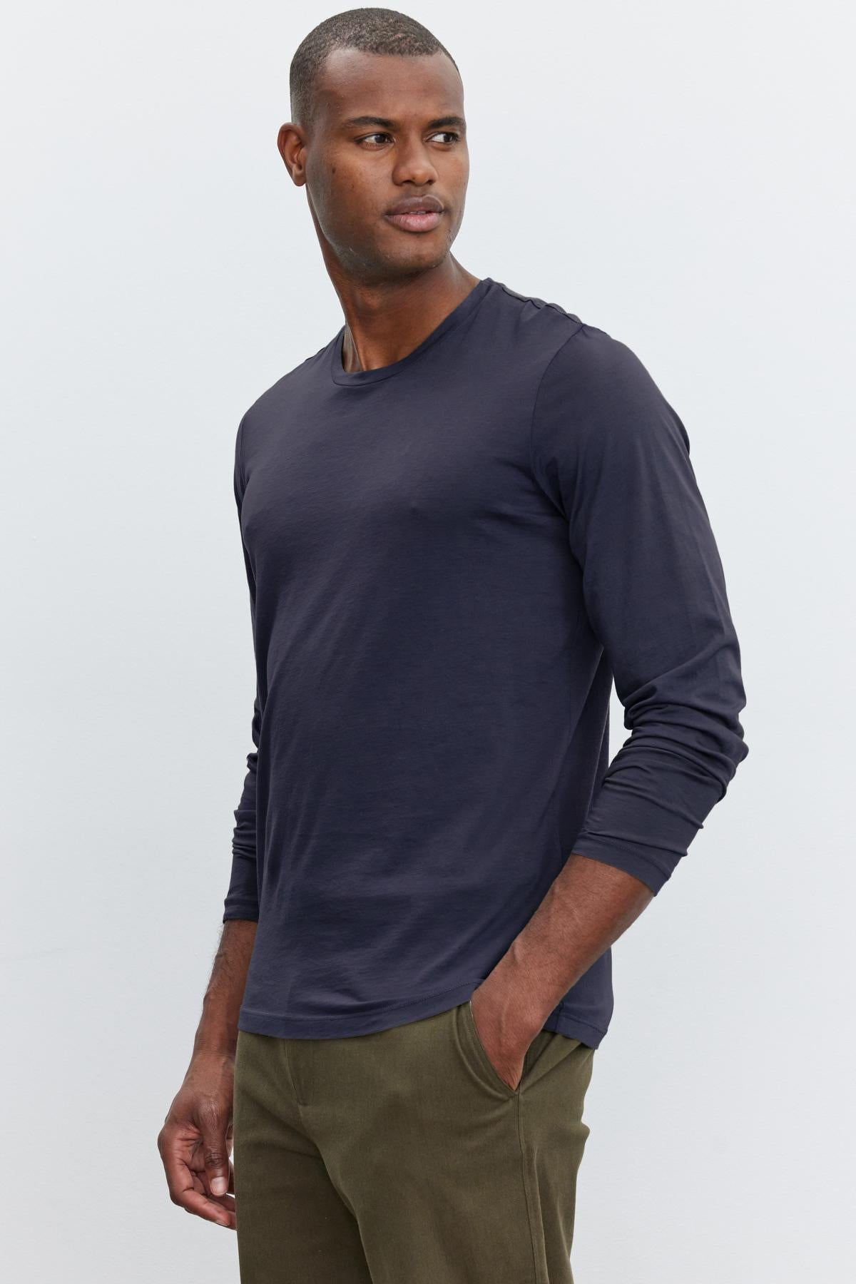   A man in a Velvet by Graham & Spencer Skeeter Whisper Classic Crew Neck Tee and olive pants stands against a plain background, looking to the side. 