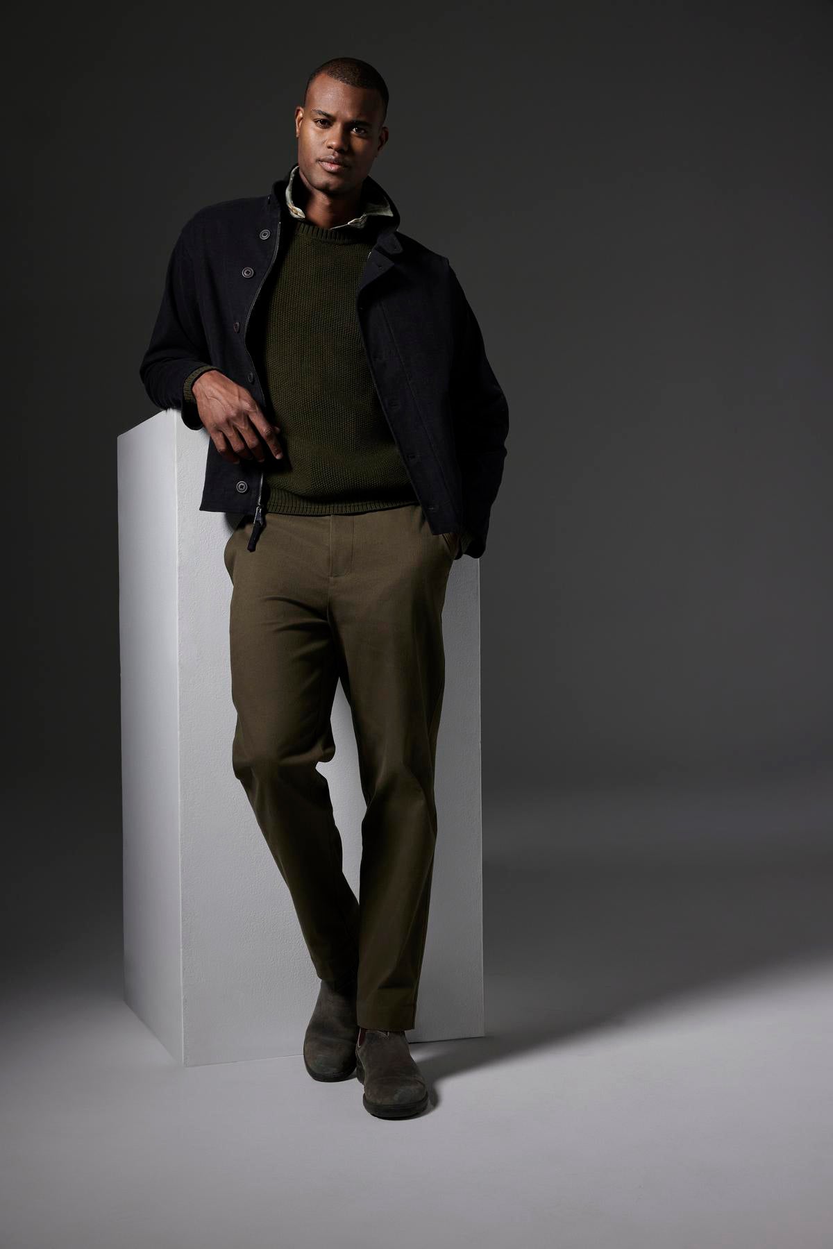   A person stands casually with one hand on a white block and the other in their pocket, wearing the WILBUR JACKET from Velvet by Graham & Spencer—a dark jacket with a button front—paired with a green sweater and green brushed cotton blend pants against a dark background. 