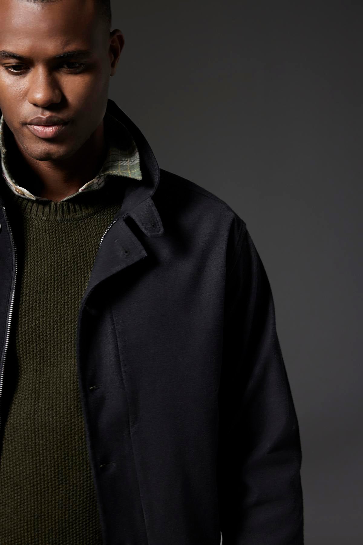   A man with short hair wearing the Velvet by Graham & Spencer WILBUR JACKET over a green sweater and a checkered shirt stands against a dark, plain background, his ensemble featuring a brushed cotton blend texture that adds depth to his look. 