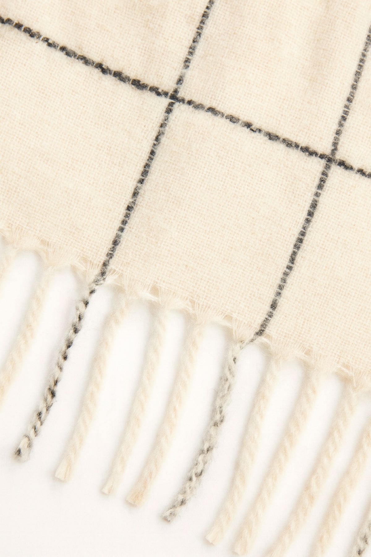 Close-up of the Velvet by Graham & Spencer WINDOWPANE MUFFLER, a cream-colored wool scarf featuring a black windowpane pattern and twisted fringe ends.