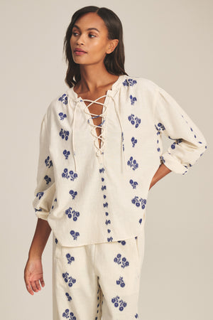 A woman wears the WINSLET TOP by Velvet by Graham & Spencer—a relaxed, boxy, long-sleeve cotton jacquard top with blue floral patterns and lace-up neckline—paired with matching pants, standing with one hand on her hip.