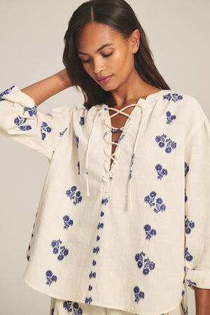 A woman wearing the WINSLET TOP by Velvet by Graham & Spencer—cream-colored with blue floral prints, long sleeves, cotton jacquard fabric, and a lace-up neckline—stands with her hand touching her hair.