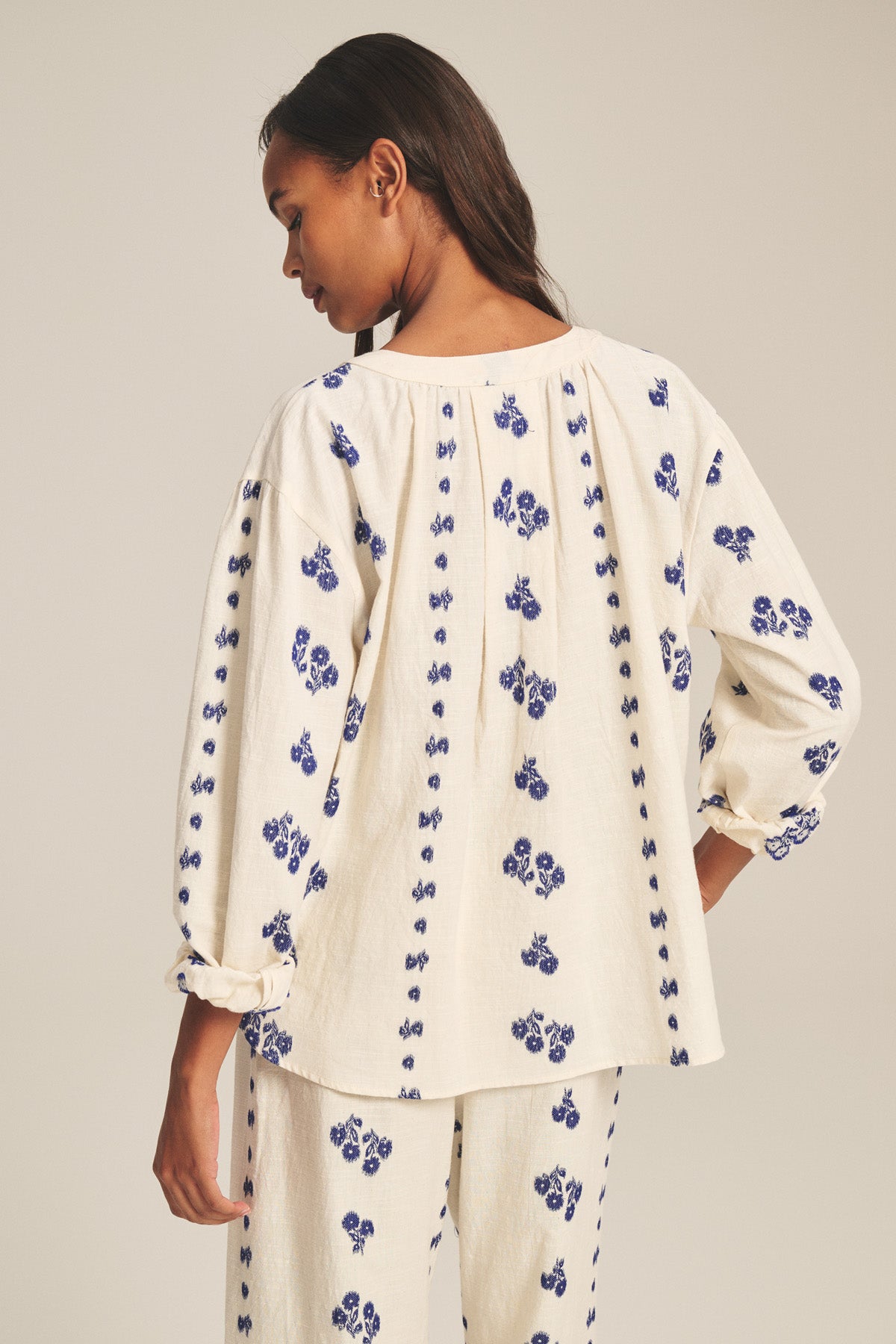 A woman faces away, wearing the WINSLET TOP by Velvet by Graham & Spencer—a relaxed boxy cotton jacquard top with blue floral patterns—paired with matching pants, against a plain beige background.-41338495959233