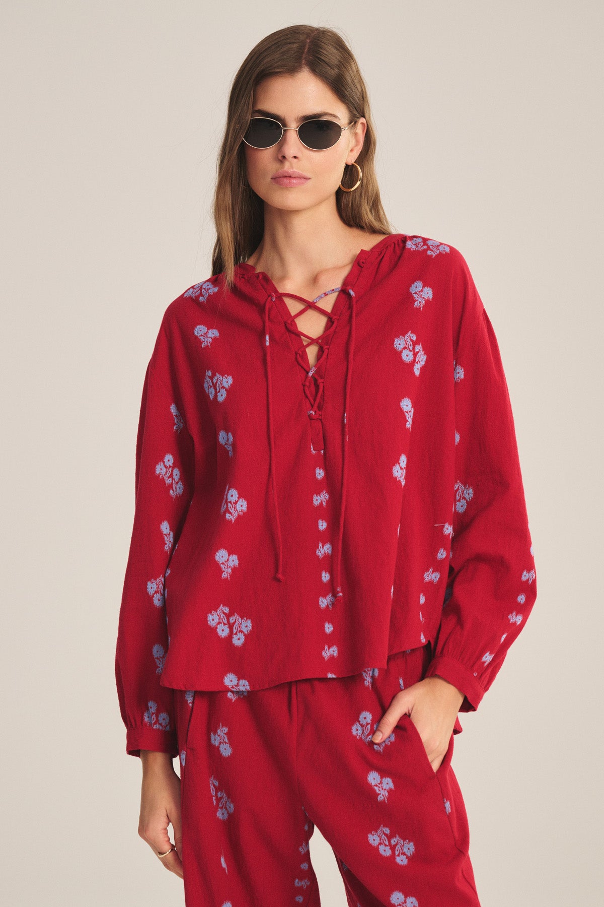 A woman in sunglasses and hoop earrings wears the Velvet by Graham & Spencer WINSLET TOP, a relaxed fit cotton jacquard with lace-up detail and small light blue florals, paired with matching red pants, standing against a plain background.-41089090093249