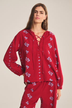 A woman wears the relaxed-fit WINSLET TOP by Velvet by Graham & Spencer— a red long-sleeve cotton jacquard piece with small blue floral patterns and matching pants— while standing against a plain background.