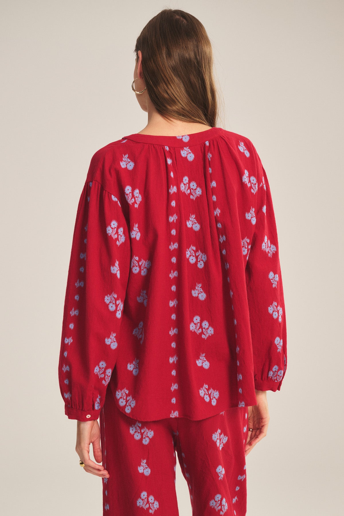 Woman with long brown hair faces away, wearing the WINSLET TOP by Velvet by Graham & Spencer—a relaxed fit red cotton jacquard top with blue floral patterns and matching pants—standing against a neutral background.