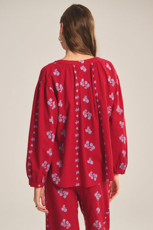 Woman with long brown hair faces away, wearing the WINSLET TOP by Velvet by Graham & Spencer—a relaxed fit red cotton jacquard top with blue floral patterns and matching pants—standing against a neutral background.