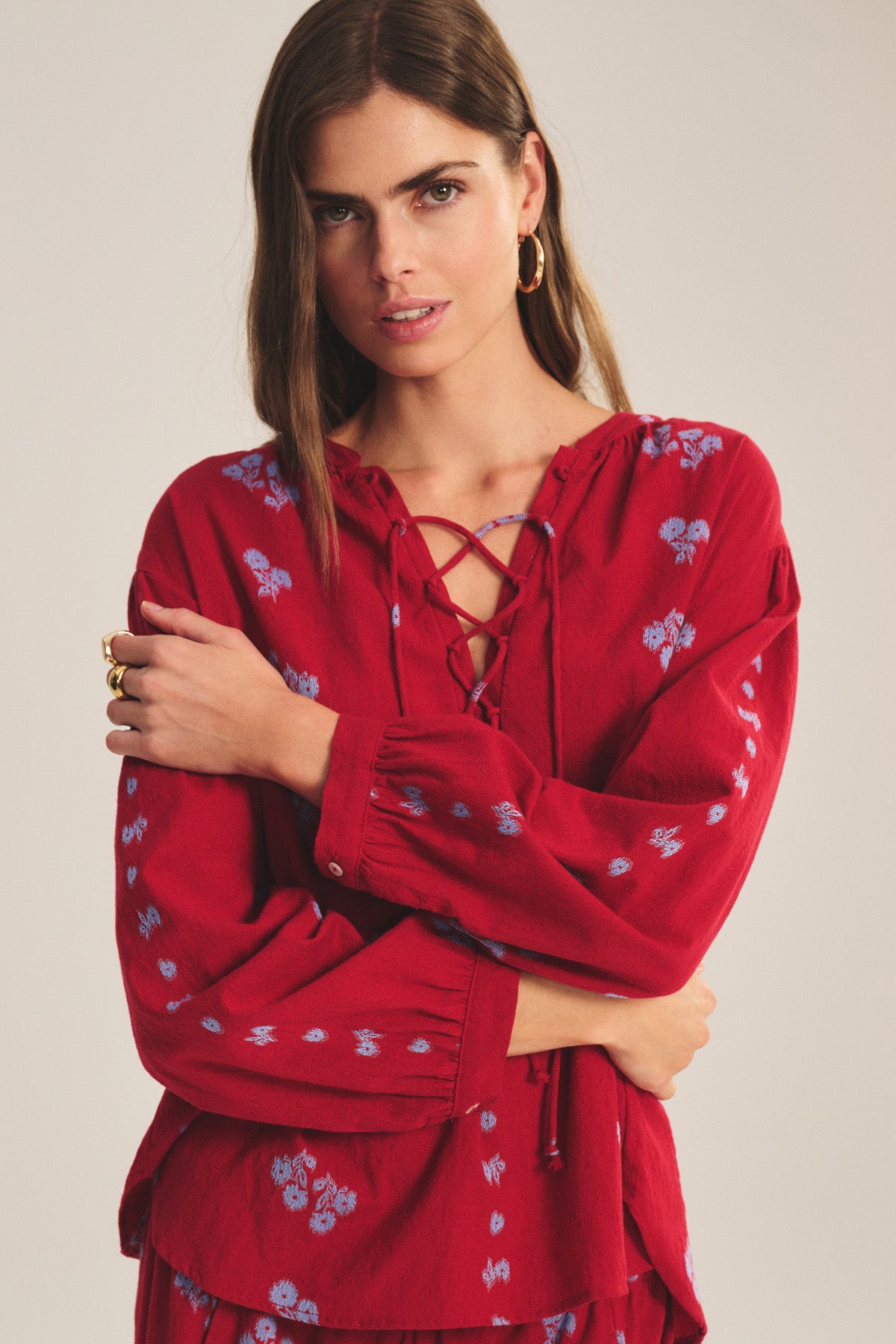 A woman stands with arms crossed wearing the WINSLET TOP by Velvet by Graham & Spencer, a red cotton jacquard top with blue floral patterns and lace-up neckline detail.-41089089929409