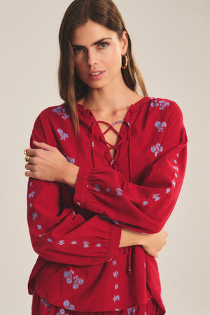 A woman stands with arms crossed wearing the WINSLET TOP by Velvet by Graham & Spencer, a red cotton jacquard top with blue floral patterns and lace-up neckline detail.