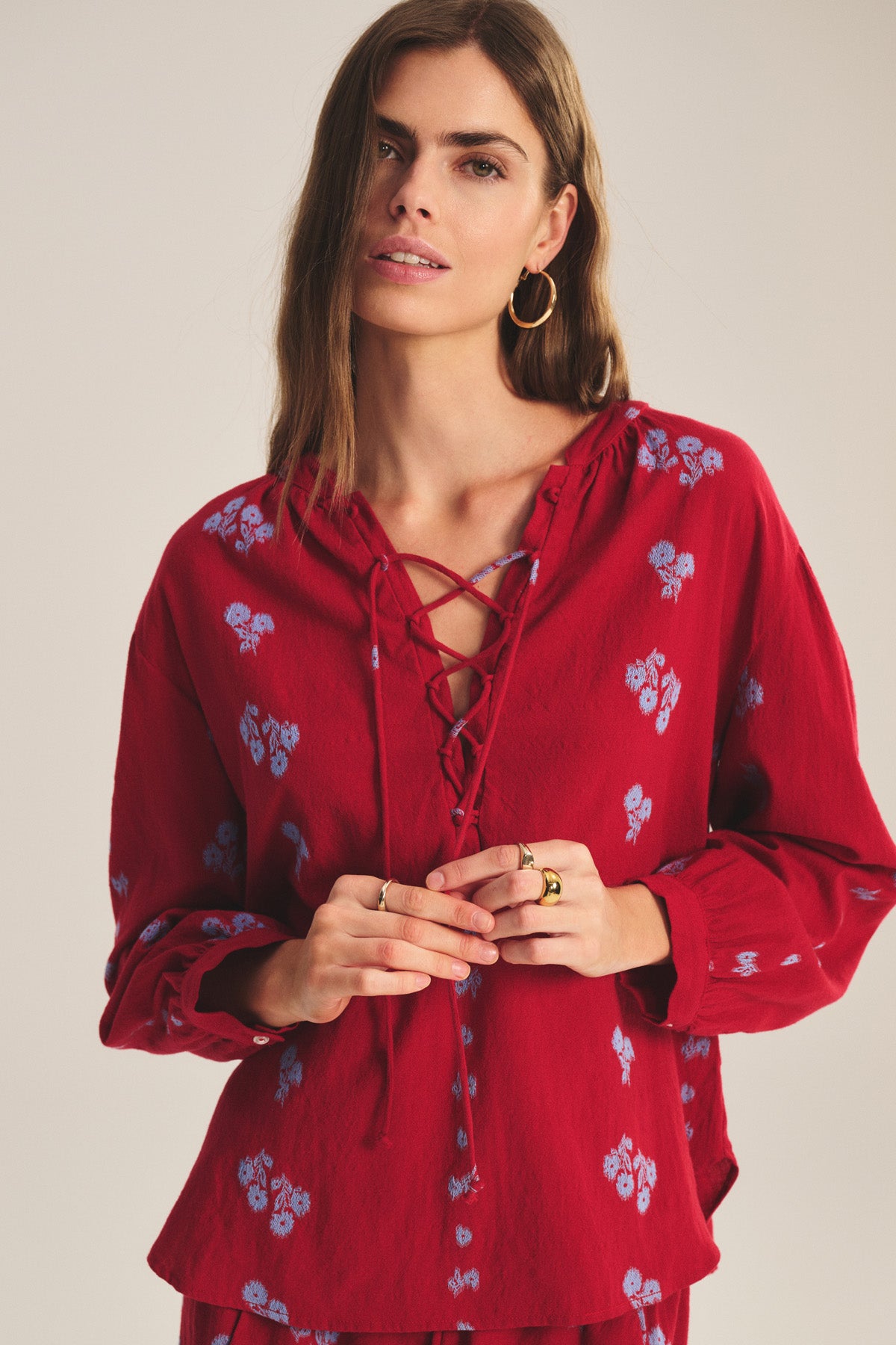 A woman stands against a neutral background wearing the WINSLET TOP by Velvet by Graham & Spencer, a red relaxed fit cotton jacquard top featuring blue floral patterns and a lace-up neckline.-41089089896641