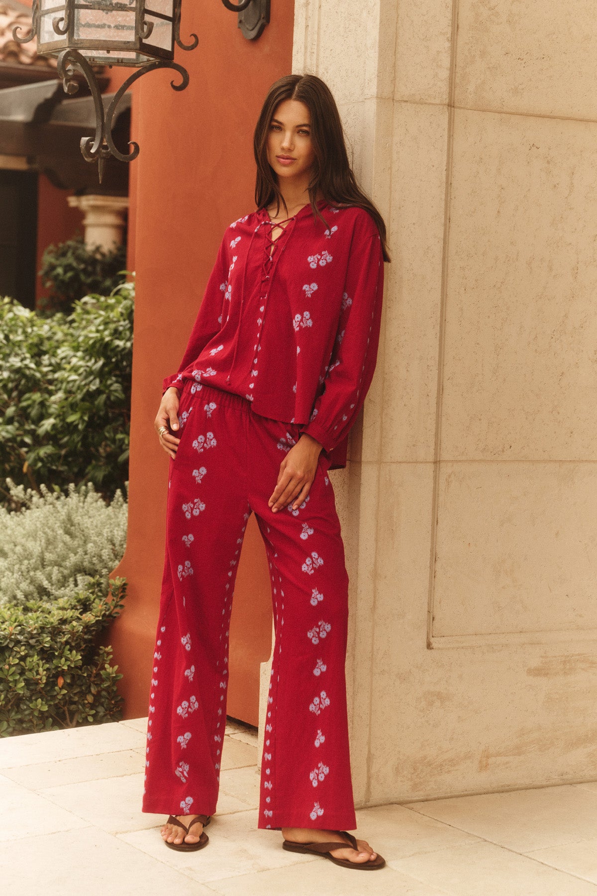 A woman models the NELLIE PANT by Velvet by Graham & Spencer: a red pajama-style set with blue floral patterns and wide-leg pants, standing outdoors against a beige stone wall with greenery and a lamp in the background.-41089042251969