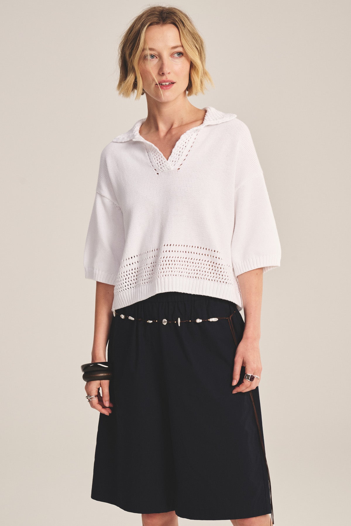 A woman stands against a plain background wearing the WINSLOW SWEATER POLO by Velvet by Graham & Spencer—a white cropped sweater polo with pointelle detailing—paired with a black skirt, shell belt, bracelets, and a ring.